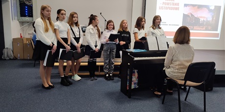 Powiększ grafikę: Chór szkolny w trakcie śpiewu- dziewczęta stoją przy pianinie, nauczycielka muzyki Anna Bazylewicz gra na pianinie.