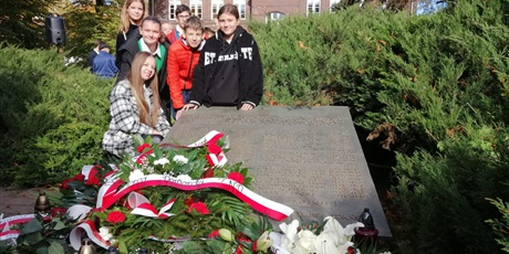 Powiększ grafikę: Uczniowie stoją przy płycie pomnika. N a pomniku leżą wieńce z kwiatami 