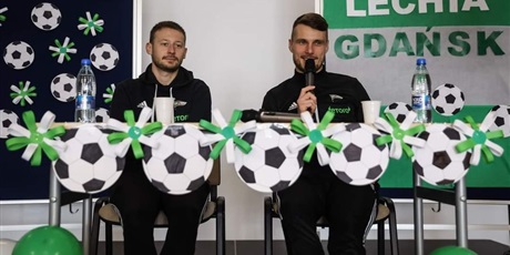 Powiększ grafikę: Dwóch zawodników Lechii Gdańsk przy stole z mikrofonami w ręku. Stół ozdobiony dekoracją z piłek w kolorach klubu