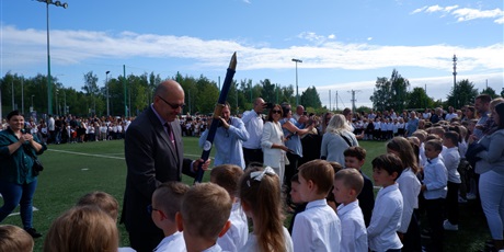 Powiększ grafikę: Uczniowie klas pierwszych  w strojach galowych stoją na boisku podczas pasowania na ucznia. Dyrektor trzyma symboliczne pióro i pasuje uczniów.